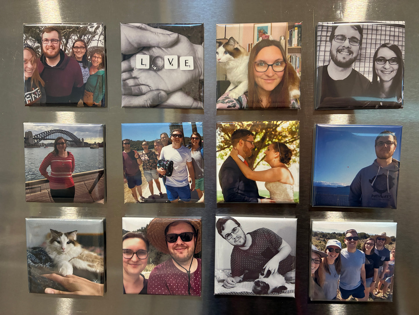A set of square custom photo magnets arranged in a 3x4 grid on a metallic silver refrigerator door. The magnets feature various family, pet, and travel photographs.