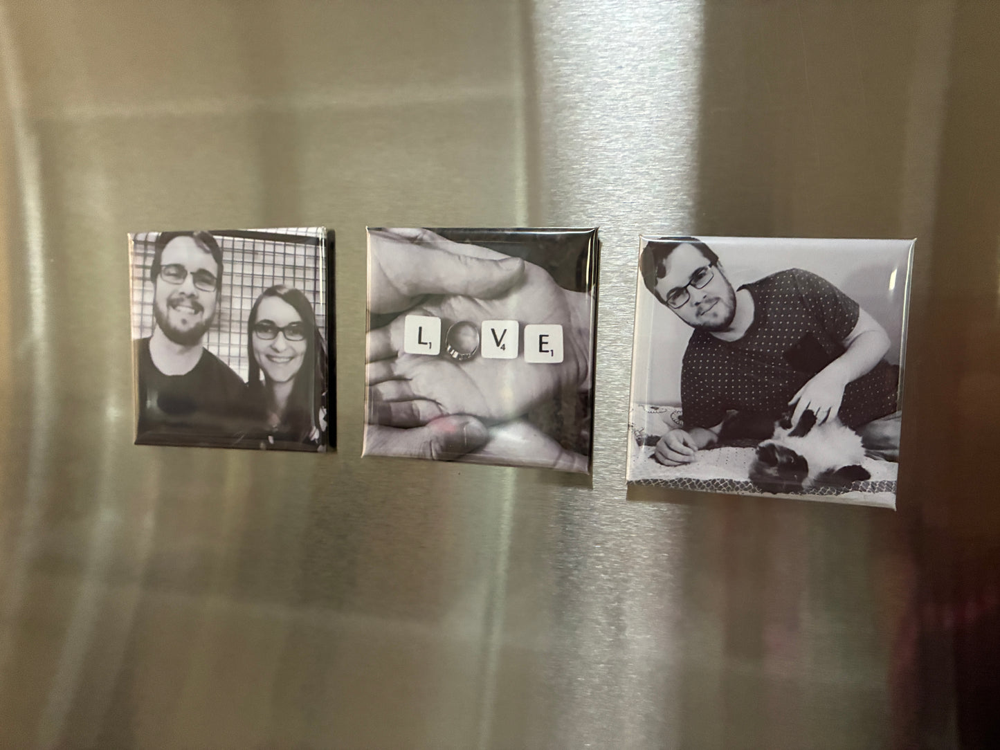Three square custom photo magnets displayed on a brushed stainless steel refrigerator door. The left magnet shows a black and white couple portrait, the middle magnet features a hand with letter tiles spelling 'LOVE', and an engagement ring as the 'O' and the right magnet shows a black and white portrait of a man petting a cat.