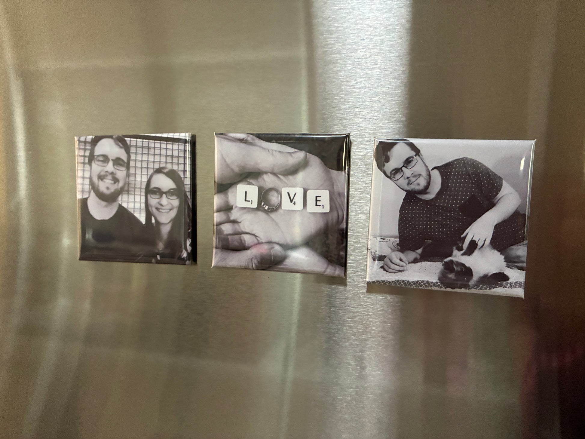 Three square custom photo magnets displayed on a brushed stainless steel refrigerator door. The left magnet shows a black and white couple portrait, the middle magnet features a hand with letter tiles spelling 'LOVE', and an engagement ring as the 'O' and the right magnet shows a black and white portrait of a man petting a cat.