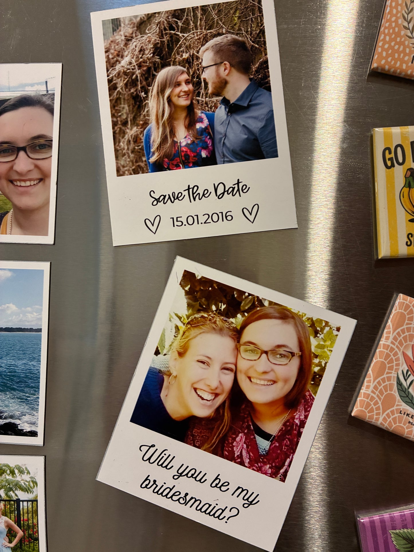 Two custom Polaroid-style photo magnets with text on a stainless steel refrigerator, surrounded by other magnets and gift boxes.