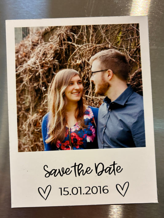 A custom instant Polaroid-style 'Save the Date' magnet on a stainless steel refrigerator. The magnet features a printed photo of a smiling couple, with the text 'Save the Date'
