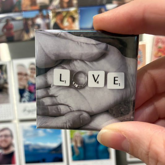 A hand holds a square magnet featuring a black and white photo of hands with scrabble letters 'L', 'V', 'E' and an engagement ring as the 'O', spelling 'LOVE'. The background shows several other magnets on a fridge